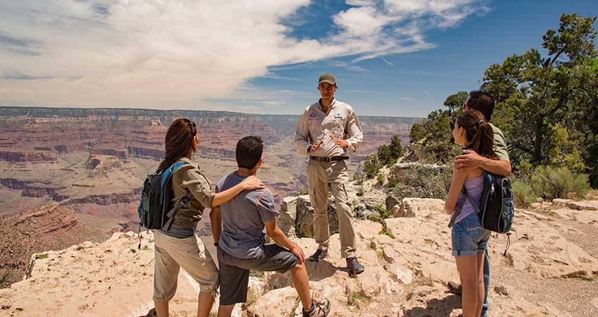 Pink Jeep Tours' Grand Canyon adventure guide talks with Grand Entrance Sunset tour guests while standing at the edge of the South Rim, with the canyon in the background.