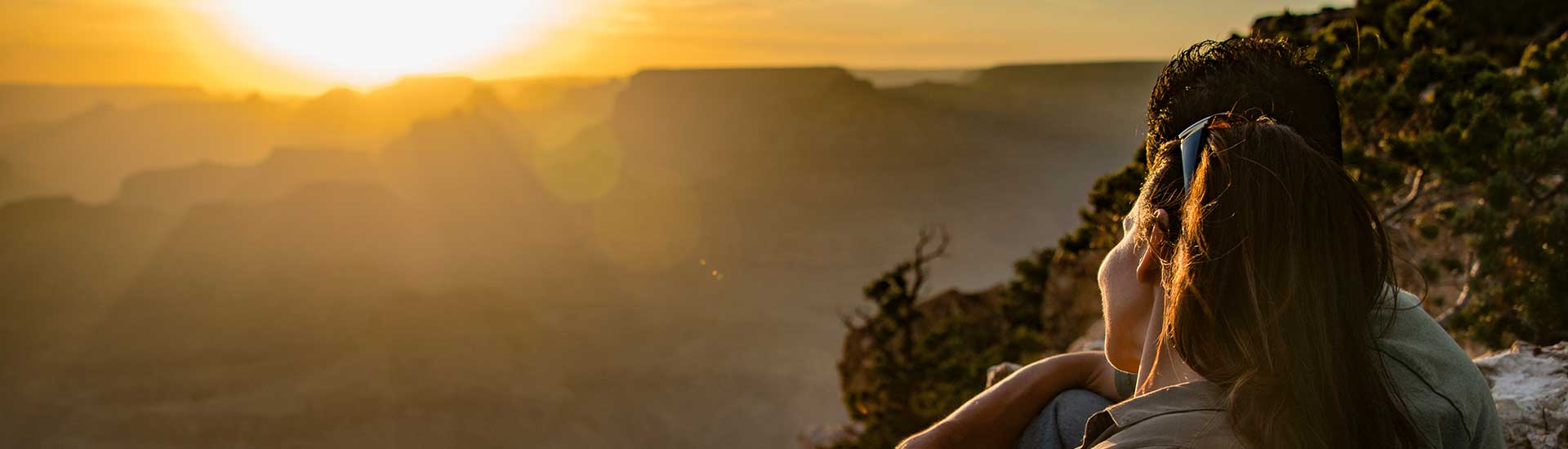 Girl looking out at a golden sun setting behind the jagged rock formations along the Grand Canyon's South Rim.