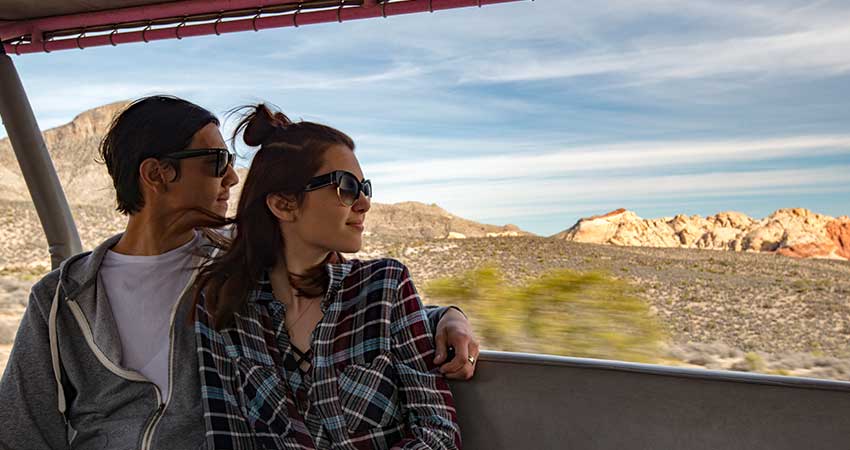 Young couple seated in the back of an open-air Pink Jeep Wrangler panoramic view of Red Rock Canyon scenery in the background.