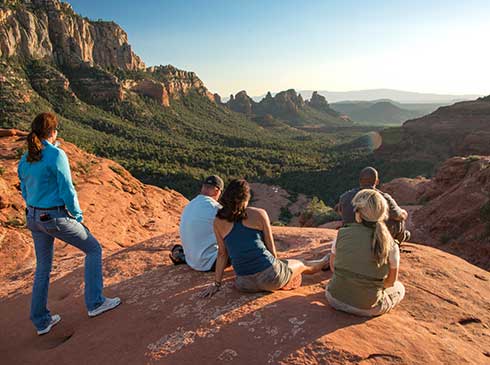 Four tour guests seated atop red rock vista point viewing Sedona and Verde Valley area below