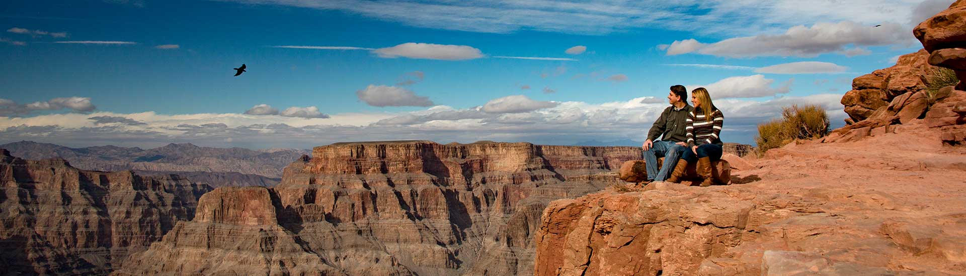 A couple seated on a rock ledge overlooking the Grand Canyon West Rim with a large bird flying above, agains a deep blue sky.