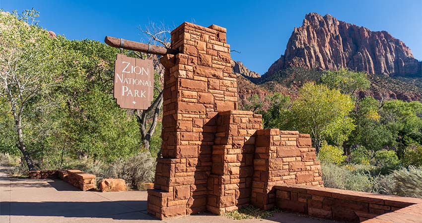 Zion National Park sign at the South Entrance pedestrian walkway with The Watchman sandstone mountain summit in the background.