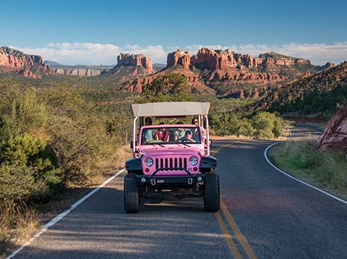 Pink Jeep Wrangler driving toward camera along Sedona's Upper Red Rock Loop road with Cathedral Rock landscape in the background, 