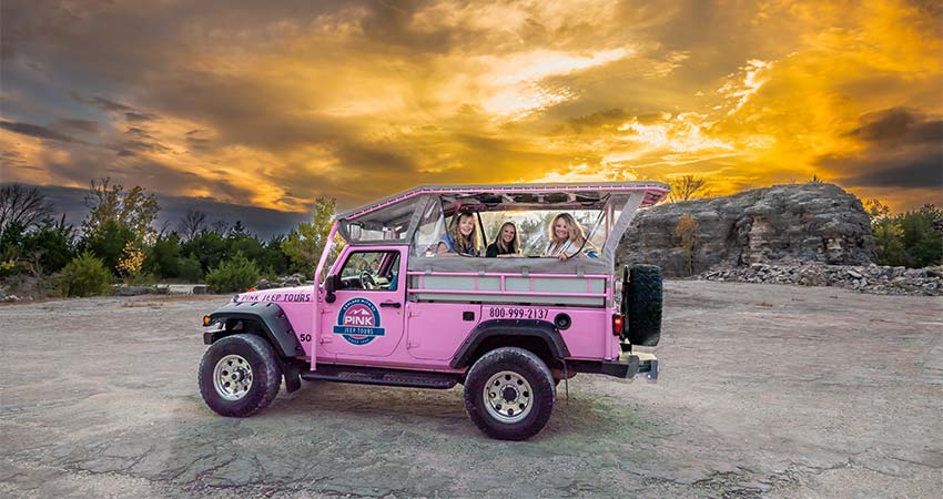 Three ladies looking out from the back of an open-air Pink Jeep Wrangler at the Branson Rock Quarry, with a fiery golden sunset above.