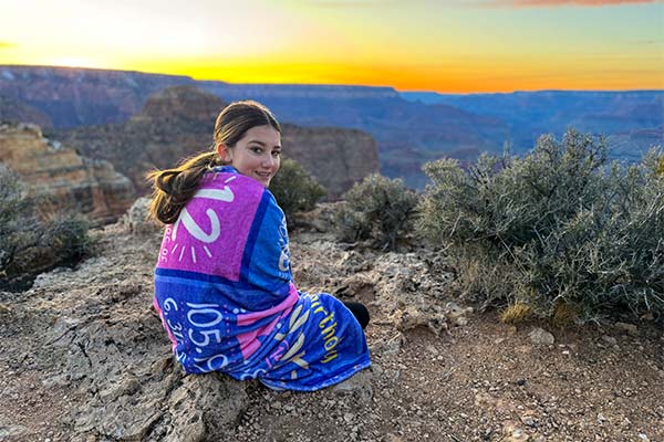 A teen girl, seated and wrapped in a colorful blanket, is facing a warm sunset over the Grand Canyon South Rim, looking back at the camera on a Pink Jeep Tour.