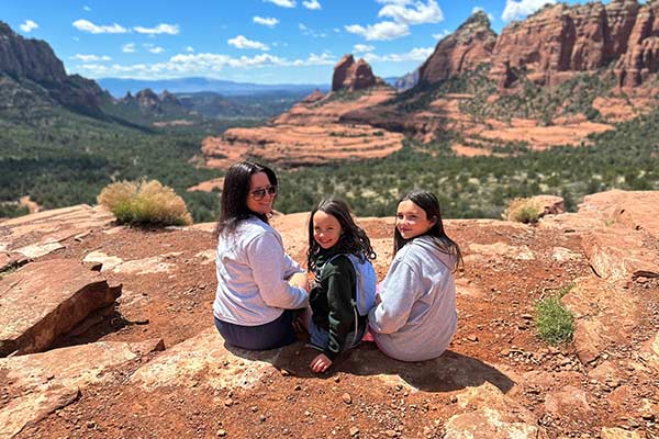 Mother and two young girls sitting on a rocky ledge facing the Cow Pies overlooking Sedona, AZ, and looking back at the camera, Pink Jeep Tours Scenic Rim tour.