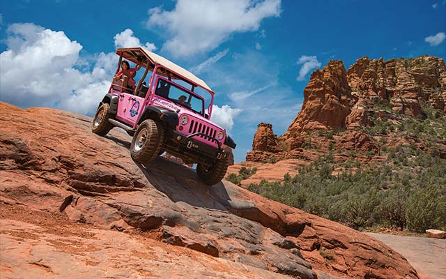 Pink Jeep Wrangler perched atop the steep red rock formations on the Broken Arrow Trail with tour guests holding onto the back seat rails.