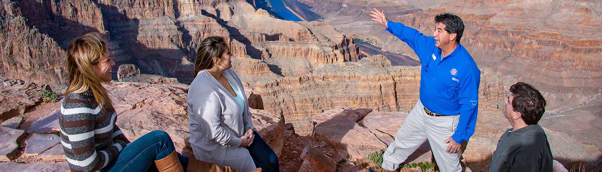 Pink Jeep Tours Grand Canyon guide talking with three guests at the edge of the Grand Canyon West Rim and pointing toward the Colorado River below.