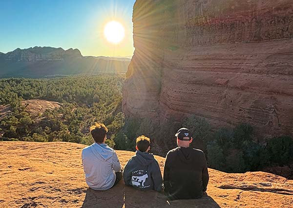 A family of three sits on a sandstone slickrock ledge at sunset, watching a golden orb dip towards the horizon casting sunrays over  Sedona's red rock landscape.