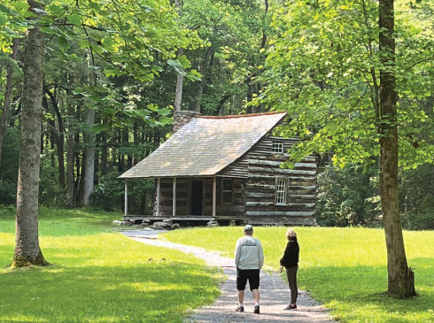 A couple looks back toward a sun-dappled log cabin in Cades Cove, Great Smoky Mountains National Park, TN.