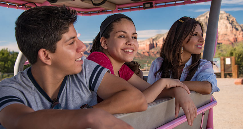 Close-up of a mother and two teens smiling while looking out from the back of a Pink Jeep Wrangler with Thunder Mountain in the background, Sedona, AZ.