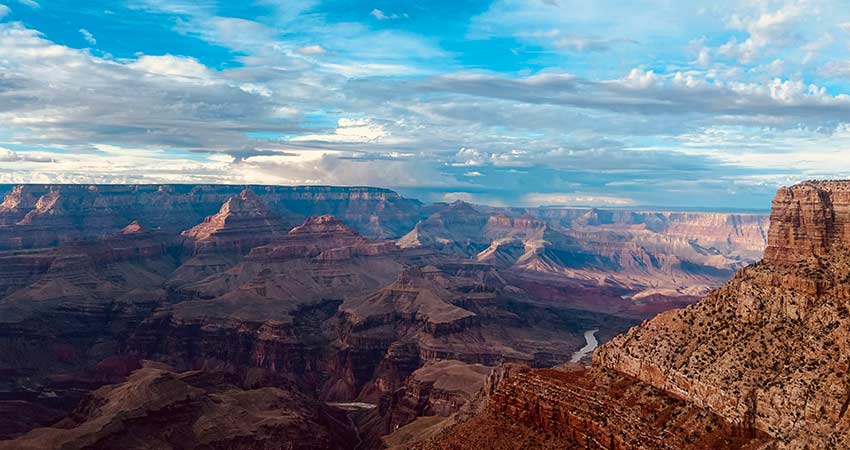 Vast Grand Canyon landscape under a blue sky with scattered clouds, showcasing layered rock formations and the Colorado River below.