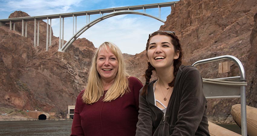 Two ladies smiling at the cameral while seated in a pontoon boat on the Colorado River below Hoover Dam, with the Mike O'Callaghan-Pat Tillman Bridge overhead.