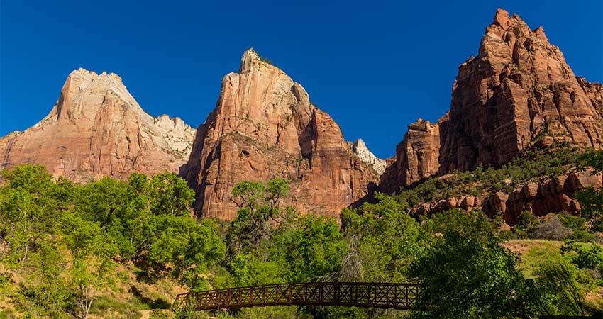 The Court of the Patriarchs tower against a deep blue sky with a bridge crossing the Virgin River and lush greenery in the foreground, Zion National Park.
