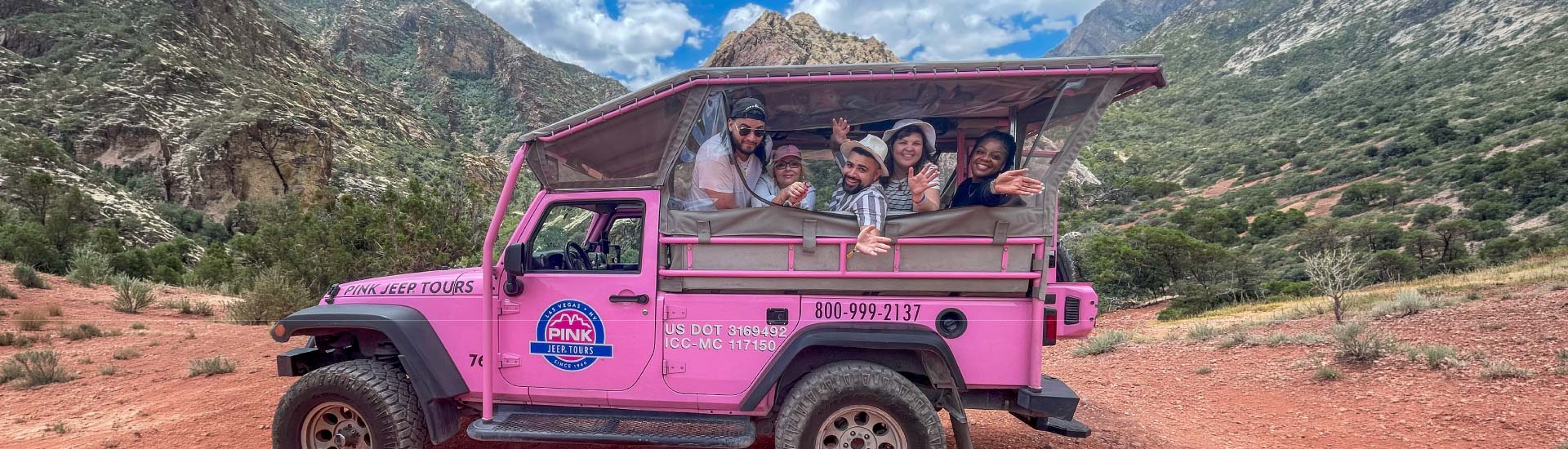 A group of friends smile from the back of a Pink Jeep in Red Rock Canyon, the mountains towering behind them