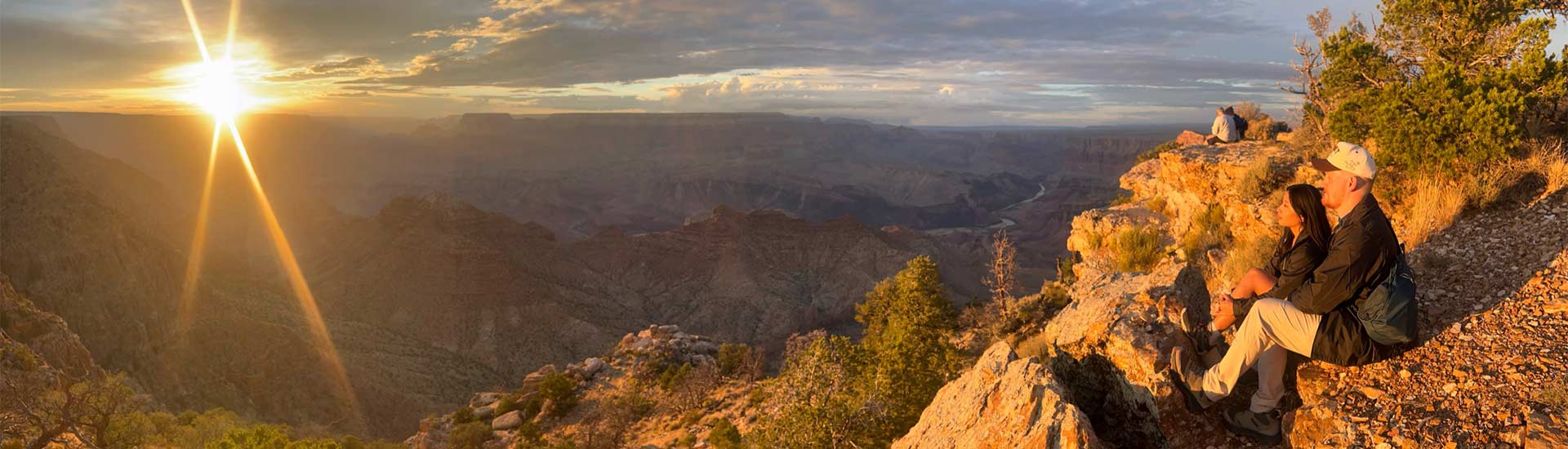 A couple sits on a rocky ledge watching a vibrant yellow sunset over the vast Grand Canyon landscape, surrounded by sparse vegetation.