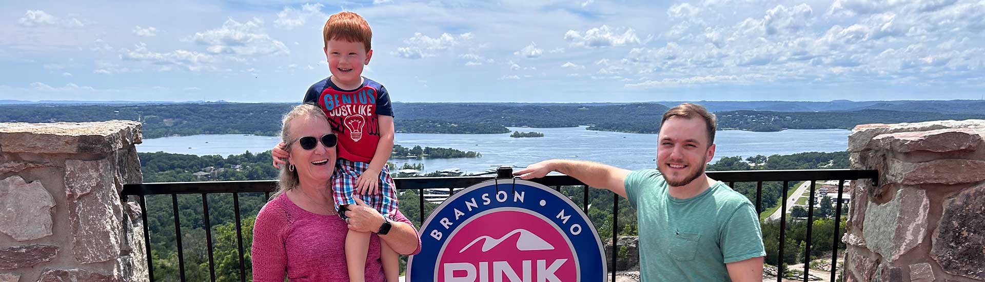 Parents and cute redheaded boy seated on mom's shoulder posing by the Pink Jeep Tours Branson, MO sign on the Baird Mountain patio, overlooking Table Rock Lake.