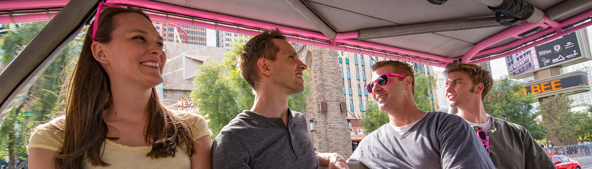 Close-up of smiling passengers riding in the back of a Pink Jeep Wrangler during the Vintage Vegas Tour, with images of the Strip in the background.