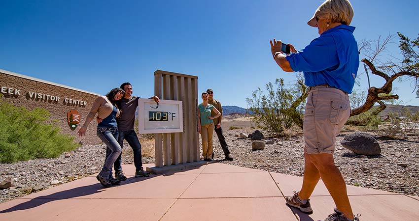 Tour guide taking couples photo at the Furnace Creek Visitor Center temperature sign reading 103 degrees, Death Valley.