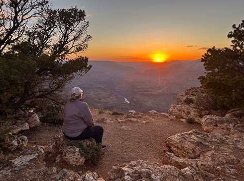 Woman seated on a boulder at the edge of the Grand Canyon watches a glowing orange sun dipping below a purple-hued horizon.