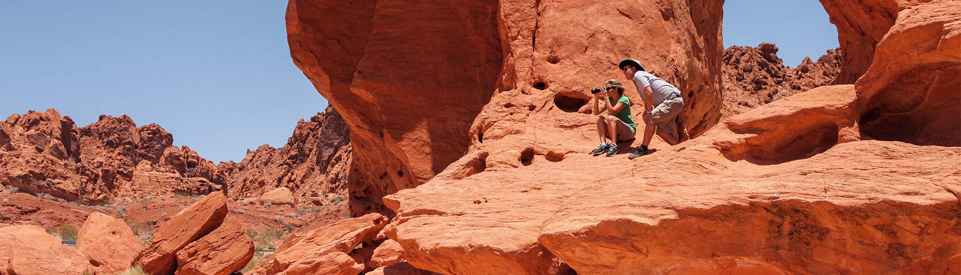 Two people sitting on the ledge of a large Aztec sandstone rock formation looking through binoculars at Valley of Fire State Park.