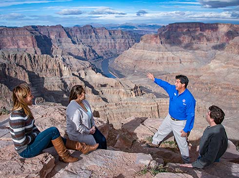 Pink Jeep Tours Grand Canyon guide kneeling beside a woman seated on a rock at the edge of the Grand Canyon's West Rim and pointing to the Colorado River below.
