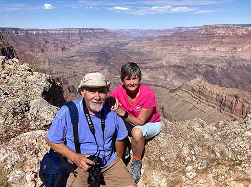 Man holding camera and woman seated on a rocky ledge facing the camera, with vast expanse of the Grand Canyon behind them.
