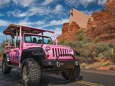 Tour guide and guests in a Pink Jeep Wrangler driving past the Chapel of the Holy Cross, built into the red rocks of Sedona, Arizona.