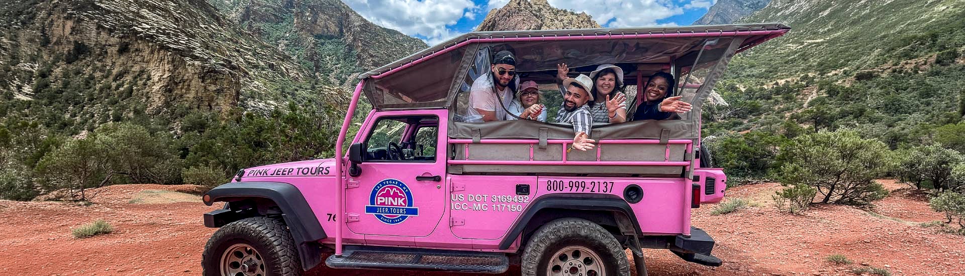 A group of friends smile from the back of a Pink Jeep in Red Rock Canyon, the mountains towering behind them