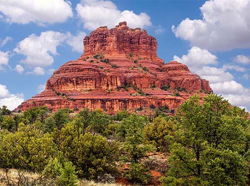 Close-up view of famous Bell Rock in Sedona Arizona, nestled against a blue sky with puffy white clouds and desert trees in the foreground.