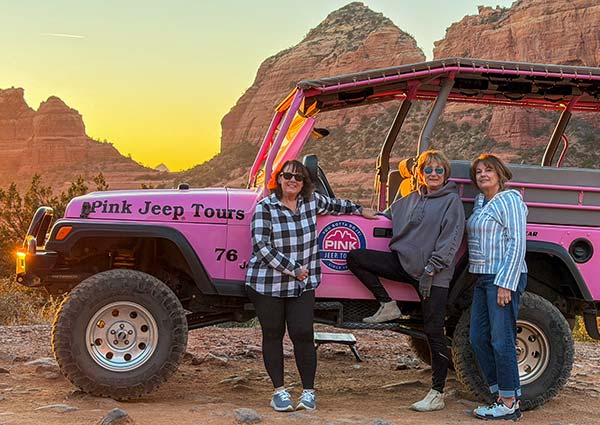 Three women leaning against a Pink Jeep Wrangler surrounded by Sedona's red rocks, with a yellow sky casting light upon them during the golden hour