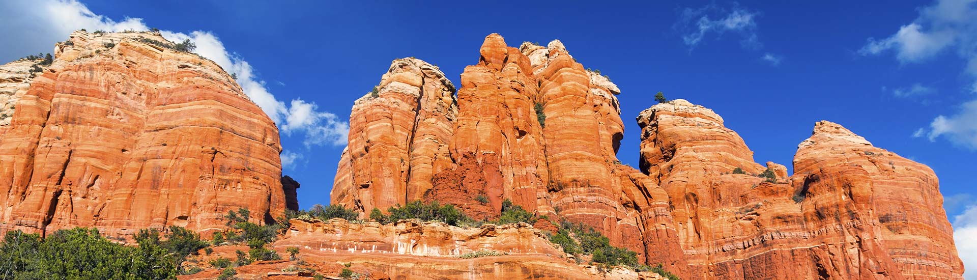 Panoramic view of Sedona's red rock formations towering against a brilliant blue sky and white clouds, Pink Jeep Tours Sedona.