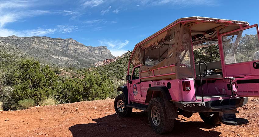 Pink Jeep Wrangler with opened back door is parked on a red dirt hill facing Turtlehead Peak at Red Rock Canyon with blue sky above.