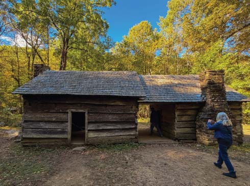 A visitor approaches a historic log cabin surrounded by fall foliage on the Roaring Fork Motor Trail in the Smoky Mountains.