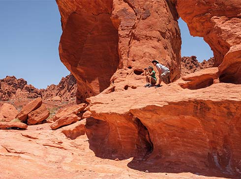 Two people sitting at the edge of a large Aztec sandstone rock formation in Valley of Fire State Park looking through binoculars.