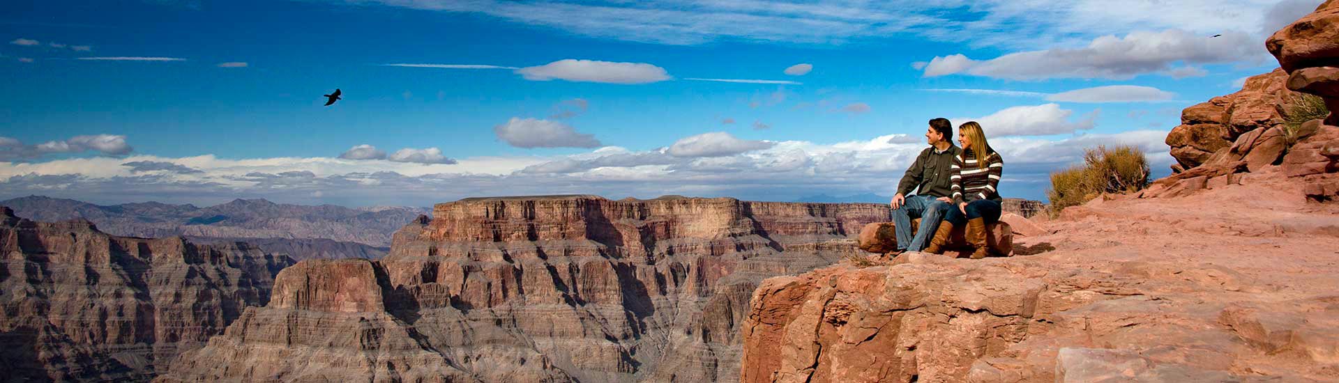 A couple seated on a rock ledge overlooking the Grand Canyon West Rim with a large bird flying above, agains a deep blue sky.