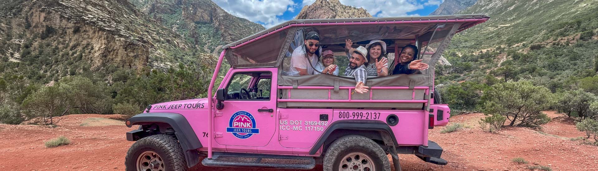 A group of friends smile from the back of a Pink Jeep in Red Rock Canyon, the mountains towering behind them