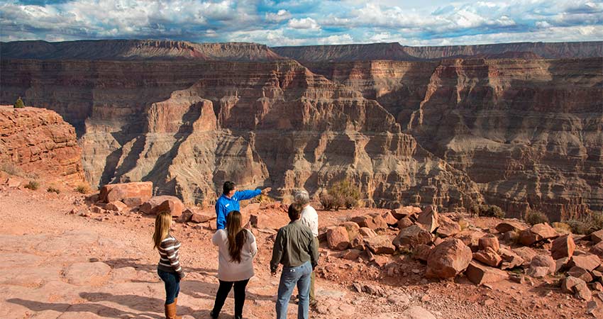 Four tour guests standing with a Pink Adventure Tours' guide at the edge of the Grand Canyon West Rim as he points towards the canyon's multicolored rock layers.