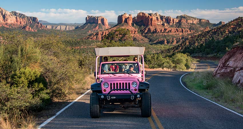 Open-air Pink Jeep Wrangler driving towards camera on Upper Red Rock Loop Road with the Cathedral Rock landscape in the background, Sedona, AZ.