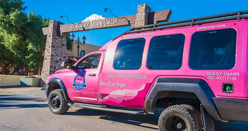 Las Vegas Pink Adventure Tour Trekker parked alongside the entrance archway of The Ranch at Death Valley with bright blue sky above.