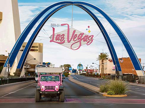 Pink Jeep Wrangler driving along the strip and passing under the Las Vegas archway with signage, Pink Jeep Tours Vintage Vegas Tour.