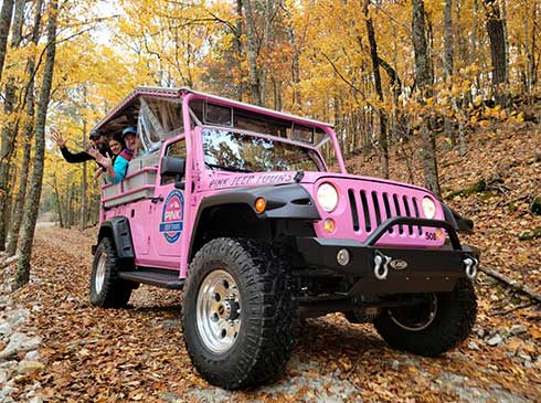 Closeup of people waving from the back of a Pink Jeep Wrangler traveling up Baird Mountain surrounded by fall leaves, Branson, MO.