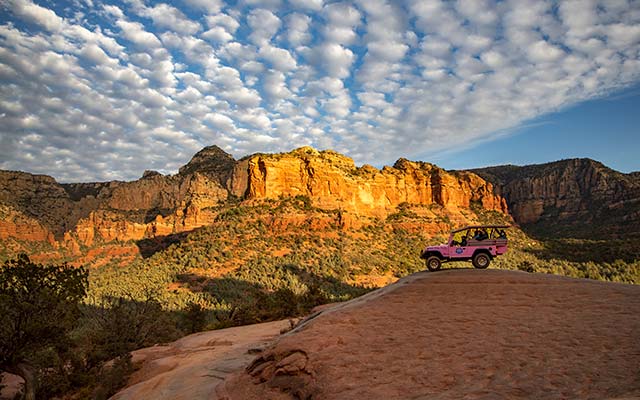 A Pink Jeep Wrangler is parked atop a sandstone slickrock mound as the sun casts a brilliant glow across Sedona's red rock landscape just before sunset.