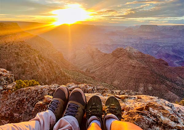 Two pairs of feet in hiking boots rest on a rocky ledge overlooking the multi-hued ridges of the Grand Canyon as a brilliant sun dips below the horizon.