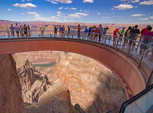 Tourists standing on the curved edged of the Grand Canyon Skywalk overlooking the Grand Canyon West Rim under a blue sky dotted with white clouds.