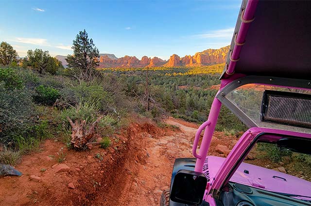 View of Sedona's red rock landscape from the driver's seat of a Pink Jeep Wrangler as it navigates a rocky off-road trail during the golden hour.