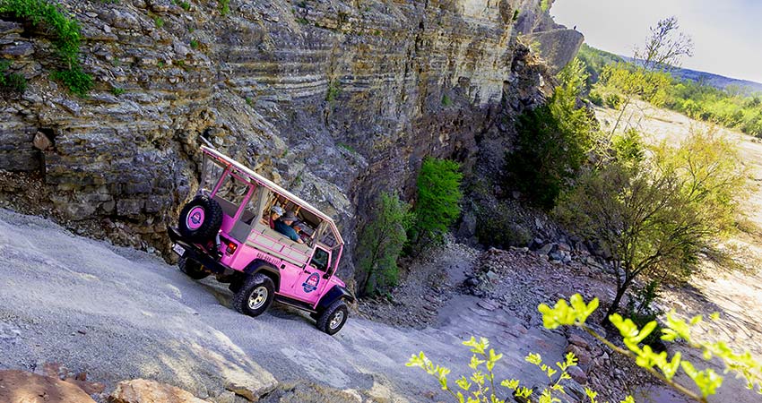 A Pink Jeep Wrangler descends an off-road, 4x4 trail along the cliff face of Baird Mountain framed by green trees and boulders.