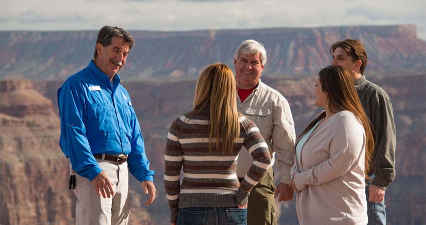 Close-up of a Pink Adventure Tours Grand Canyon adventure guide smiling and talking with four tour guests and the Grand Canyon West Rim in the background.