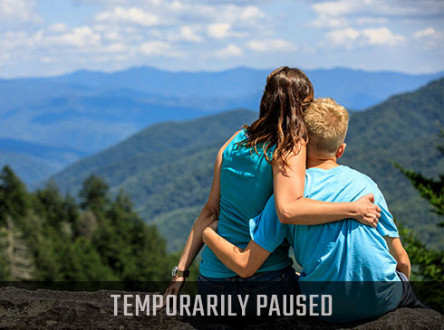 Mother and son seated on Newfound Gap Overlook wall looking towards the Smoky Mountains with Temporarily Paused Banner.