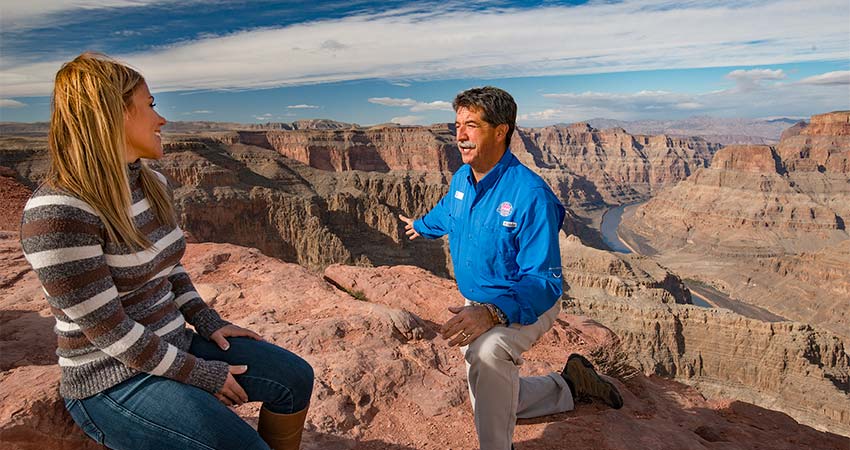 Pink Jeep Tours Grand Canyon guide kneeling beside a woman seated on a rock at the edge of the Grand Canyon's West Rim and pointing to the Colorado River below.